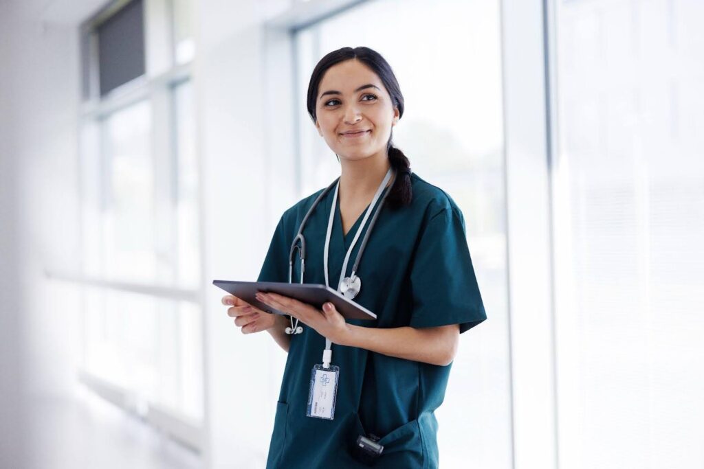 A smiling nurse standing in a medical facility corridor holds a clipboard.