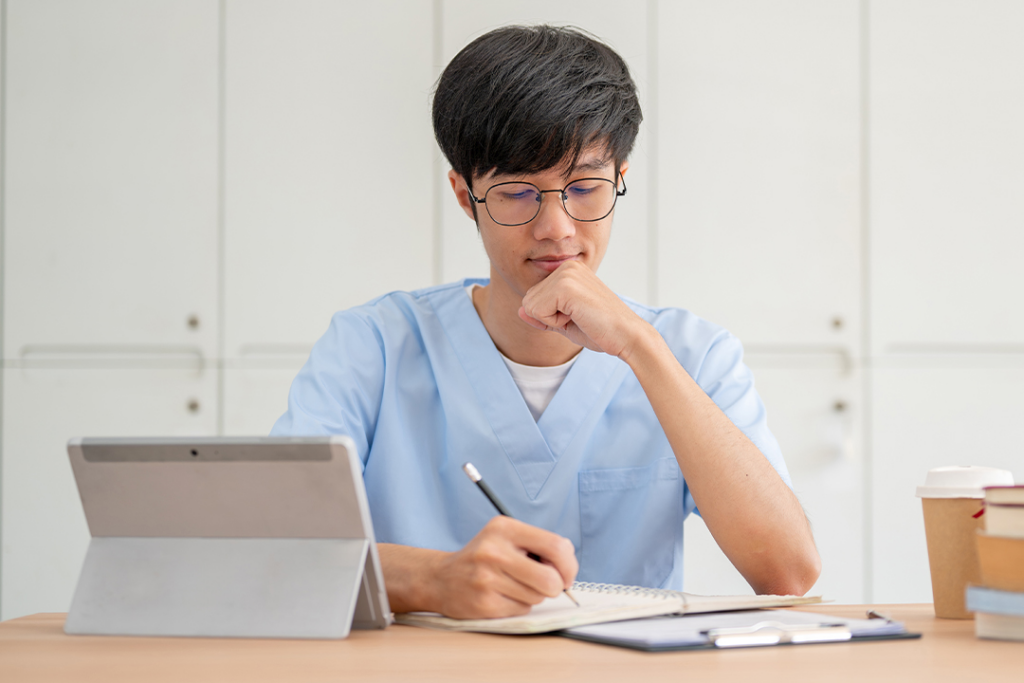 A health professional writes on a clipboard while a tablet stands upright beside him.